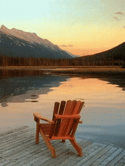 An Adirondack Chair Sits On A Dock Overlooking A Lake At Sunset GIF