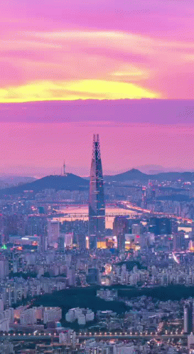 An Aerial View Of A City At Sunset With A Tall Building In The Foreground GIF