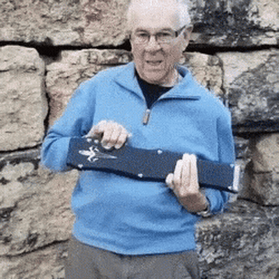An Elderly Man In A Blue Sweater Is Holding A Guitar Case In Front Of A Stone Wall . GIF