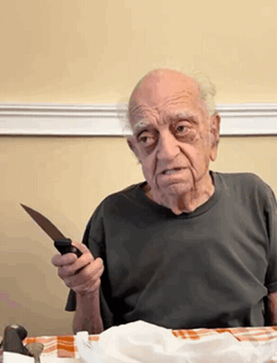 An Elderly Man Is Sitting At A Table Holding A Knife In His Hand GIF