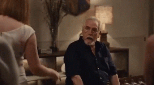 An Elderly Man Is Sitting On A Couch In A Living Room With A Woman Standing Next To Him . GIF