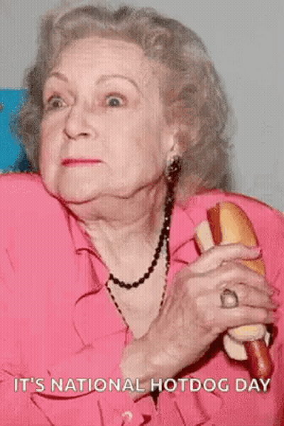 An Elderly Woman In A Pink Shirt Is Holding A Hot Dog And Says It 'S National Hotdog Day . GIF