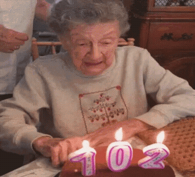 An Elderly Woman Is Blowing Out Candles On A Cake With The Number 102 On It GIF