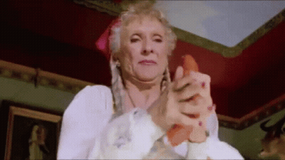 An Elderly Woman Is Holding A Carrot In Her Hand And Making A Funny Face . GIF