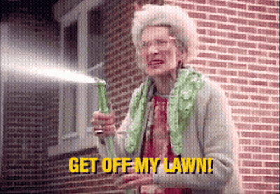 An Elderly Woman Is Holding A Hose In Front Of A Brick Building And Saying `` Get Off My Lawn '' . GIF