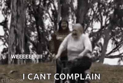 An Elderly Woman Is Riding A Bike In The Woods While A Man Looks On . GIF