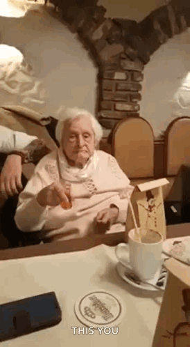 An Elderly Woman Is Sitting At A Table With A Cup Of Coffee And A Spoon . GIF