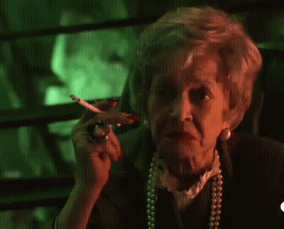An Elderly Woman Is Smoking A Cigarette And Waving At The Camera In Front Of A Green Background . GIF