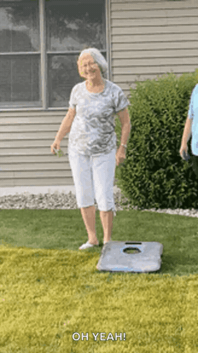 An Elderly Woman Is Standing On A Cornhole Board In A Grassy Yard . GIF