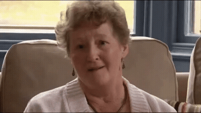 An Elderly Woman Wearing A White Sweater And Earrings Is Sitting On A Couch GIF