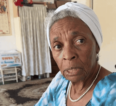 An Elderly Woman Wearing A White Turban And Pearls Looks At The Camera GIF