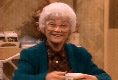 An Elderly Woman Wearing Glasses And A Blue Sweater Is Holding A Cup Of Coffee And Smiling . GIF