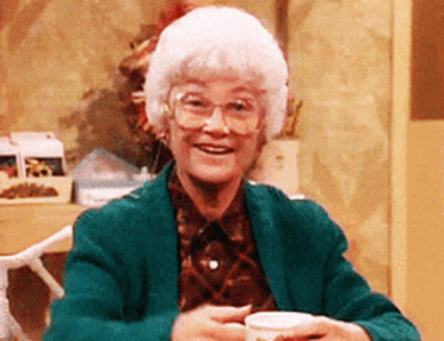 An Elderly Woman Wearing Glasses And A Green Jacket Holds A Cup Of Coffee GIF