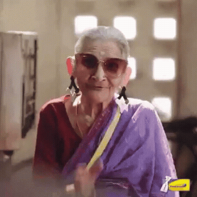 An Elderly Woman Wearing Sunglasses And A Purple Sari Is Smiling And Waving . GIF