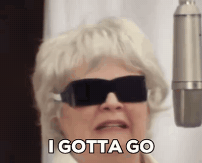 An Elderly Woman Wearing Sunglasses And A Wig Is Standing In Front Of A Microphone . GIF