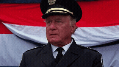 An Older Man In A Police Uniform Stands In Front Of A Flag GIF