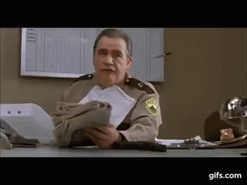 An Older Man In A Sheriff 'S Uniform Is Sitting At A Desk Holding A Bag Of Money . GIF