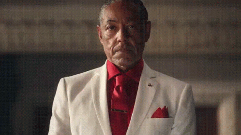 An Older Man In A White Suit And Red Tie Is Standing In A Dark Room . GIF