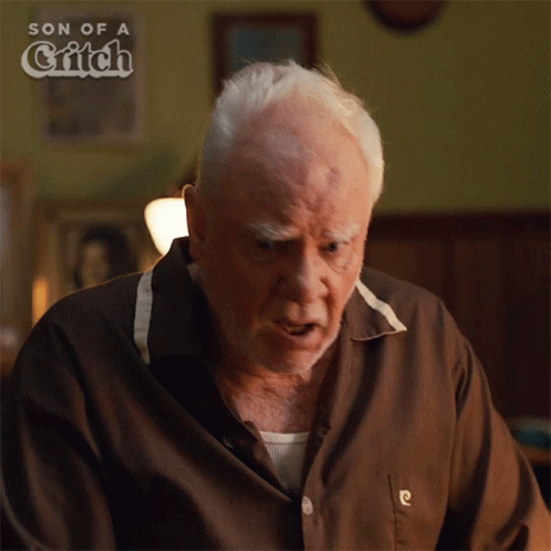 An Older Man Is Sitting In Front Of A Sign That Says Son Of A Glitch GIF