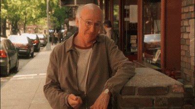 An Older Man Leans Against A Brick Wall In Front Of A Building That Says Bar GIF