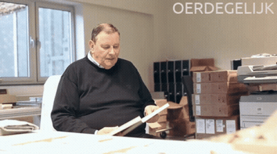 An Older Man Sits At A Desk Looking At A Book With The Word Oerdegelijk In The Corner GIF