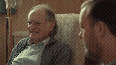 An Older Man Sitting In A Chair Talking To Another Man With The Word Gay On The Bottom GIF