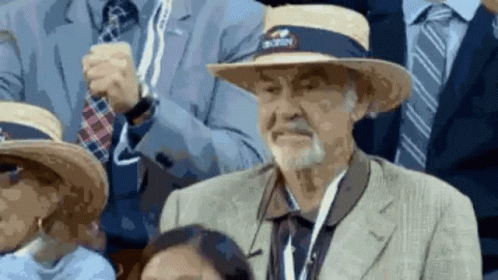 An Older Man Wearing A Straw Hat With A Nyc Logo On It Is Sitting In A Crowd Of People . GIF