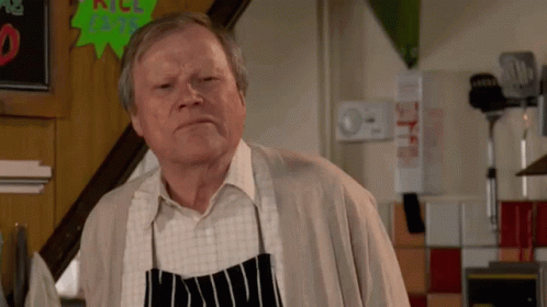 An Older Man Wearing An Apron Is Standing In Front Of A Sign That Says " All Beef " GIF