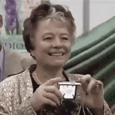 An Older Woman Is Smiling While Holding A Small Camera . GIF