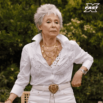 An Older Woman Wearing A White Shirt And White Pants Is Standing In Front Of A Sign That Says Fast X GIF