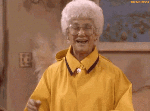 An Older Woman Wearing A Yellow Raincoat And Glasses Is Laughing GIF