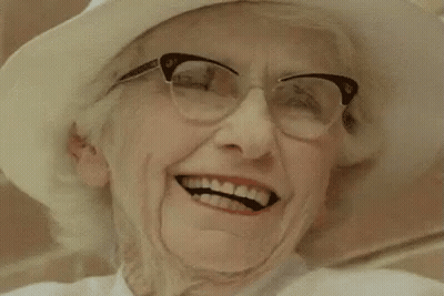 An Older Woman Wearing Glasses And A White Hat Is Smiling . GIF