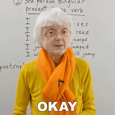 An Older Woman Wearing Glasses And An Orange Scarf Says Okay In Front Of A Whiteboard GIF