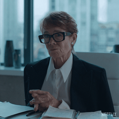 An Older Woman Wearing Glasses Sits At A Desk With A Netflix Logo On The Bottom Right GIF
