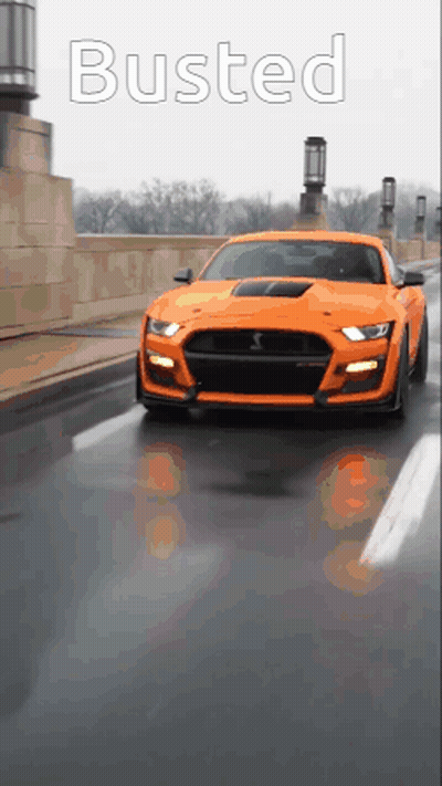 An Orange Mustang Is Driving Down A Street With The Word Busted On The Bottom GIF