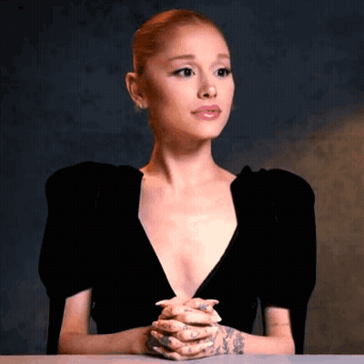 Ariana Grande Is Sitting At A Table With Her Hands Folded In Front Of Her Face . GIF
