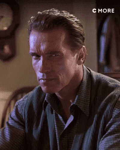 Arnold Schwarzenegger Is Sitting At A Table In Front Of A Clock And Looking At The Camera . GIF