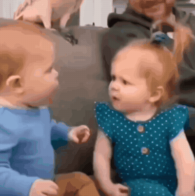 Baby And Little Girl Sitting On Couch Looking At Each Other GIF