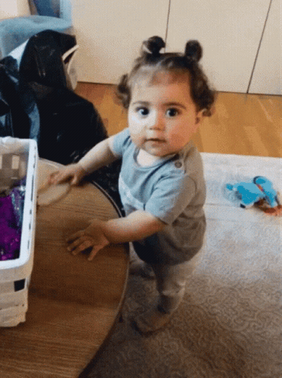 Baby Girl Standing At Wooden Table Looking At Camera GIF