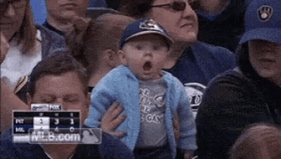 Baby In Baseball Cap Held By Woman In Crowd GIF