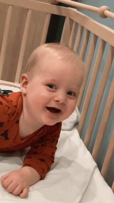 Baby In Orange Shirt Laying In Wooden Crib GIF