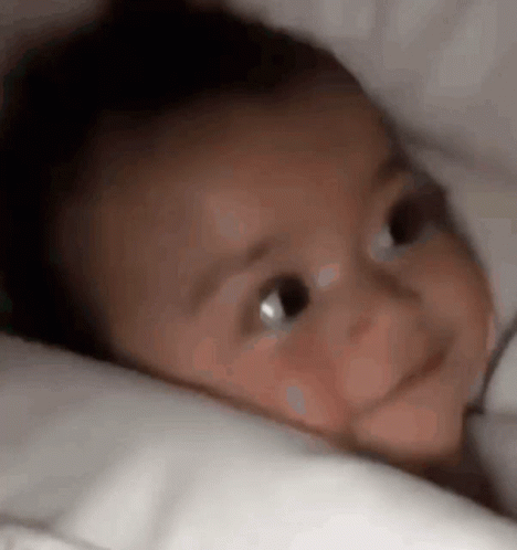 Baby Laying On Bed With White Blanket Looking At Camera GIF