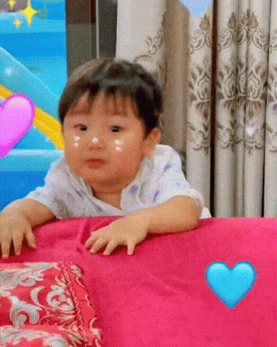 Baby Sitting On Bed With Pink Pillow And Blue Heart GIF