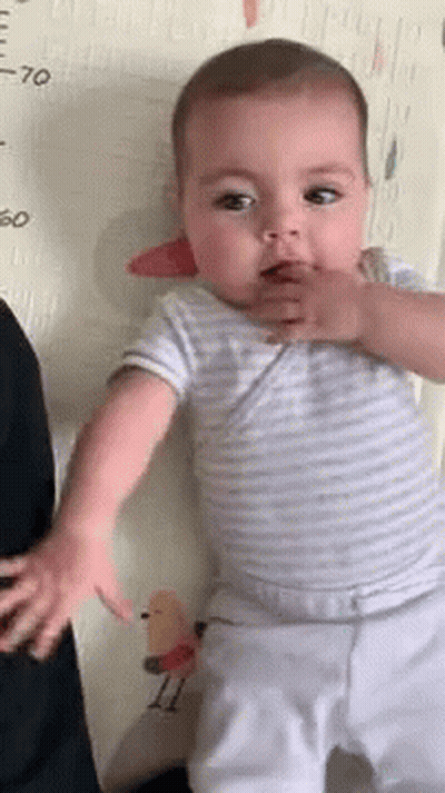Baby Standing In Front Of Measuring Tape Looking At Camera GIF