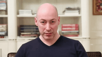Bald Man In Blue Shirt Sitting In Front Of Bookshelf GIF