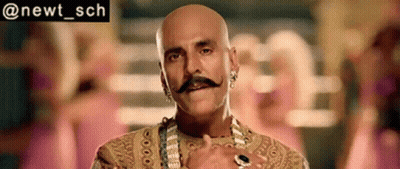 Bald Man In Gold Dress With Mustache, Earrings, And Necklace GIF