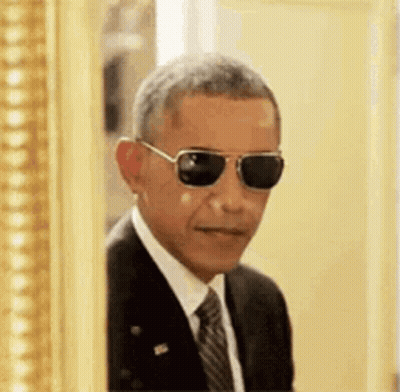 Barack Obama Is Wearing Sunglasses And A Suit And Tie While Peeking Out Of A Picture Frame . GIF