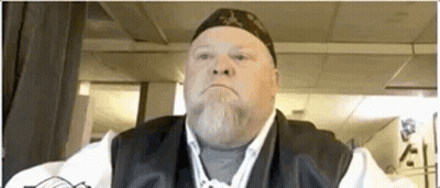 Bearded Man In Hat And Vest Looks At Camera GIF