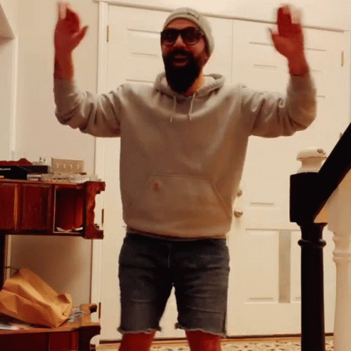 Bearded Man In Shorts And Hoodie Standing In Front Of Door With Arms In Air GIF