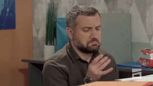 Bearded Man Sitting At Desk With Hand To Chest GIF